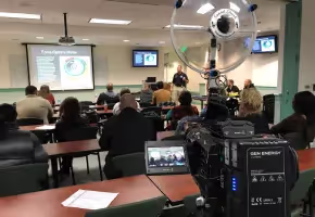 Video camera with a Sound Shark long-range microphone recording a presentation.
